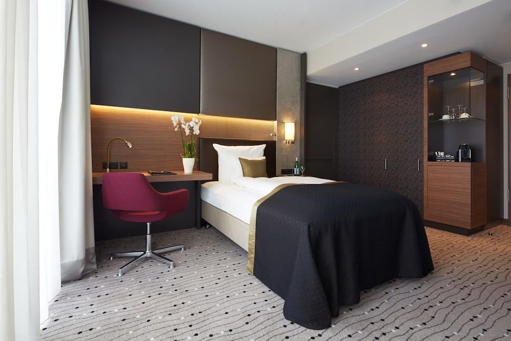Steigenberger Hotel Am Kanzleramt Βερολίνο Εξωτερικό φωτογραφία The photo shows a modern hotel room featuring a single bed with a dark bedspread. The room has a contemporary design with a neutral color palette, accented by warm lighting. There's a small desk with a red chair positioned next to the bed. A cabinet 