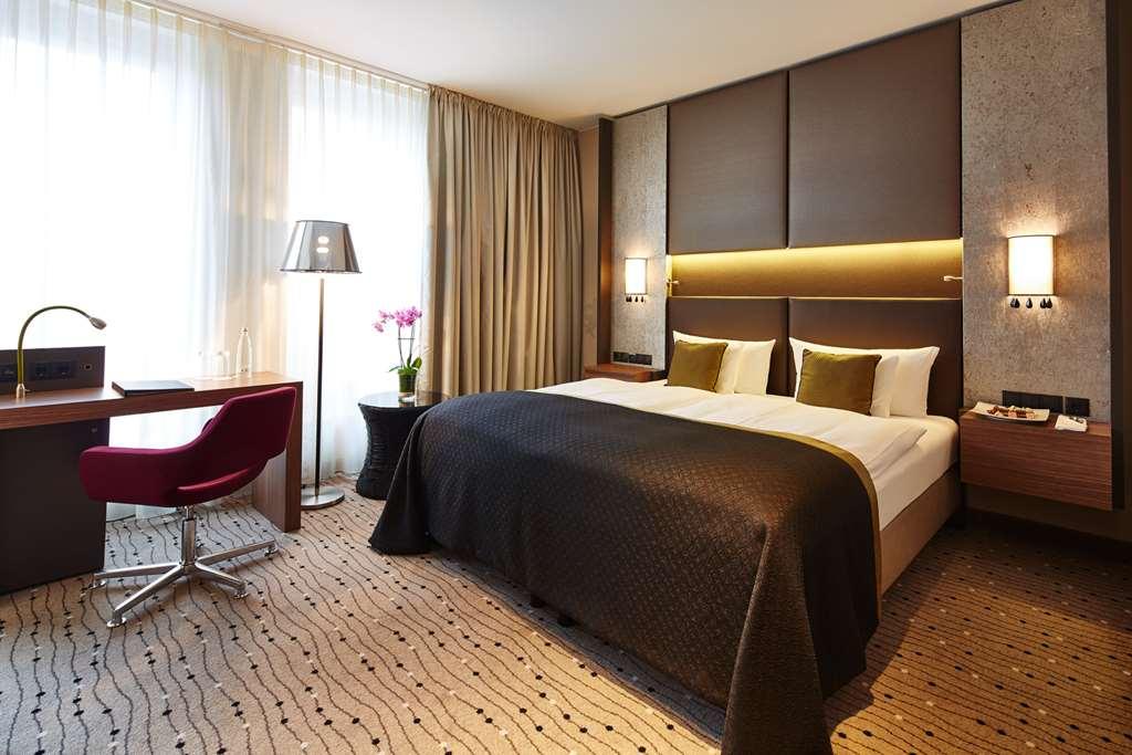 Steigenberger Hotel Am Kanzleramt Βερολίνο Δωμάτιο φωτογραφία The image shows a modern hotel room featuring a large bed with a dark bedspread and white pillows. There are two bedside lamps mounted on the wall above the bed. To one side of the room, there is a stylish desk with a red chair, and a floor lamp next
