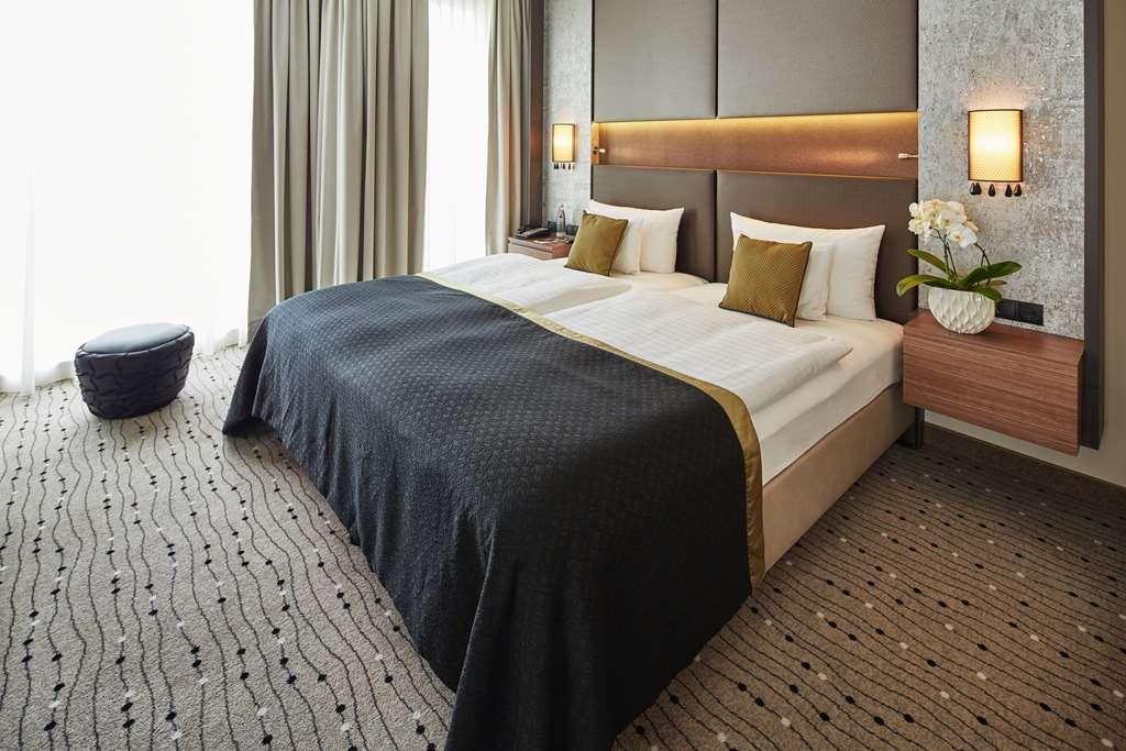 Steigenberger Hotel Am Kanzleramt Βερολίνο Δωμάτιο φωτογραφία The photo shows a modern hotel room featuring a cozy double bed with a dark coverlet and white linens. There are two pillows on each side of the bed, with one side decorated in olive green accents. On either side of the bed are stylish nightstands eq