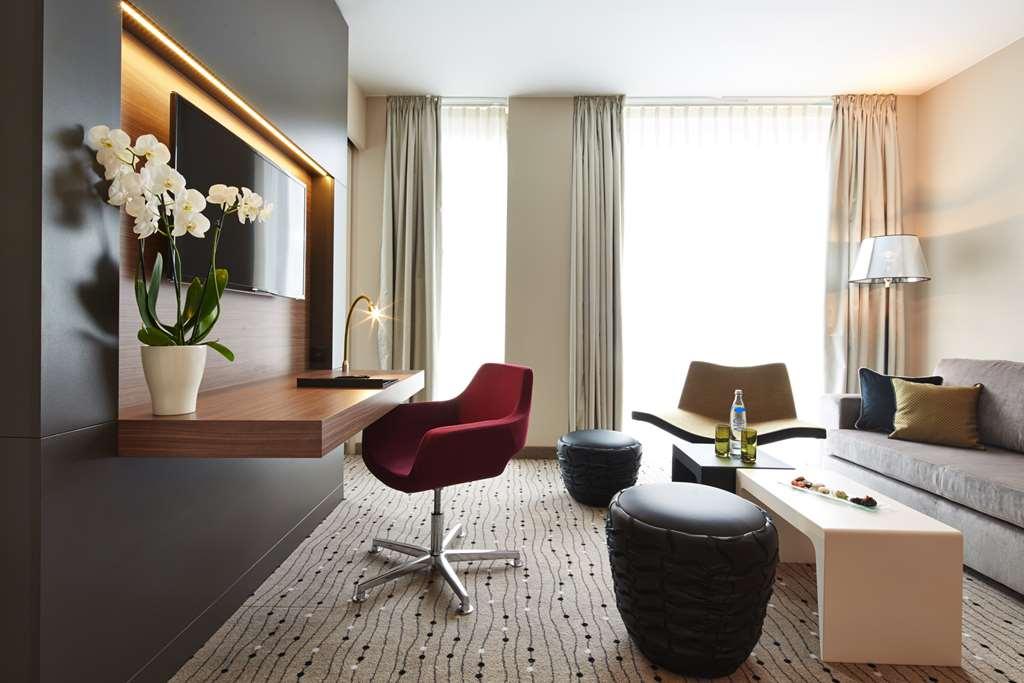 Steigenberger Hotel Am Kanzleramt Βερολίνο Δωμάτιο φωτογραφία The photo depicts a modern and stylish living area. It features a sleek design with neutral-colored walls and large windows allowing natural light to fill the space. 

Key elements include:

- A wall-mounted television.
- A stylish red chair with a s