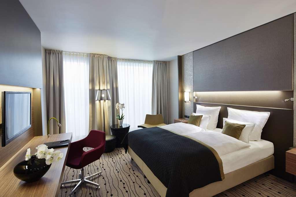 Steigenberger Hotel Am Kanzleramt Βερολίνο Δωμάτιο φωτογραφία The photo shows a modern hotel room with a minimalist design. It features a large double bed with white linens and a dark throw blanket. On either side of the bed, there are small lamps. There is a desk with a chair, and a flat-screen TV mounted on t