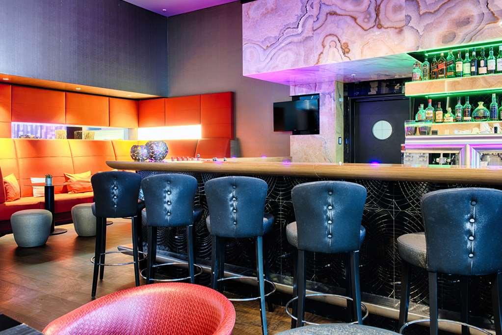 Steigenberger Hotel Am Kanzleramt Βερολίνο Εστιατόριο φωτογραφία The photo shows a modern bar area featuring a long, sleek countertop with several black upholstered barstools arranged in front of it. The bar has decorative lighting that casts a colorful glow, enhancing the ambiance. Behind the bar, there is a sele