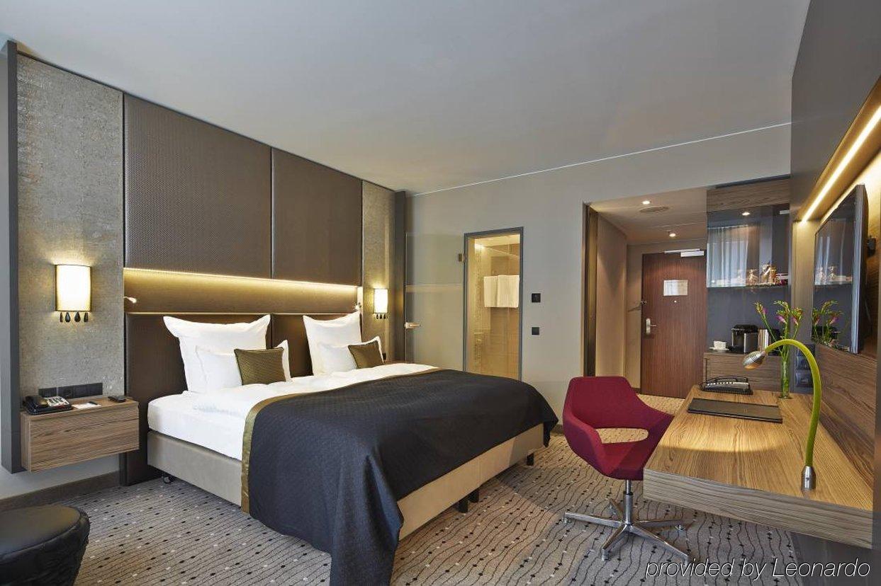 Steigenberger Hotel Am Kanzleramt Βερολίνο Εξωτερικό φωτογραφία The photo shows a modern hotel room featuring a neatly made bed with a dark-colored bedspread. There are two white pillows positioned at the head of the bed. The room has walls with a neutral color palette and stylish lighting, adding an elegant touc