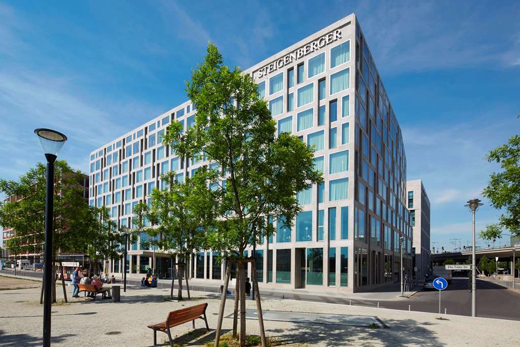 Steigenberger Hotel Am Kanzleramt Βερολίνο Εξωτερικό φωτογραφία The photo shows a modern, multi-story building with a large glass facade, featuring a grid pattern of windows. The building has the name "Steigenberger" prominently displayed at the top. Surrounding the structure are green trees and a landscaped area