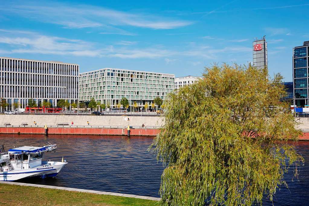 Steigenberger Hotel Am Kanzleramt Βερολίνο Εξωτερικό φωτογραφία The photo shows a modern urban landscape along a river. On one side, there are contemporary buildings with glass facades, reflecting a variety of architectural styles. In front of these structures, there is a green area with a tree, suggesting a blen