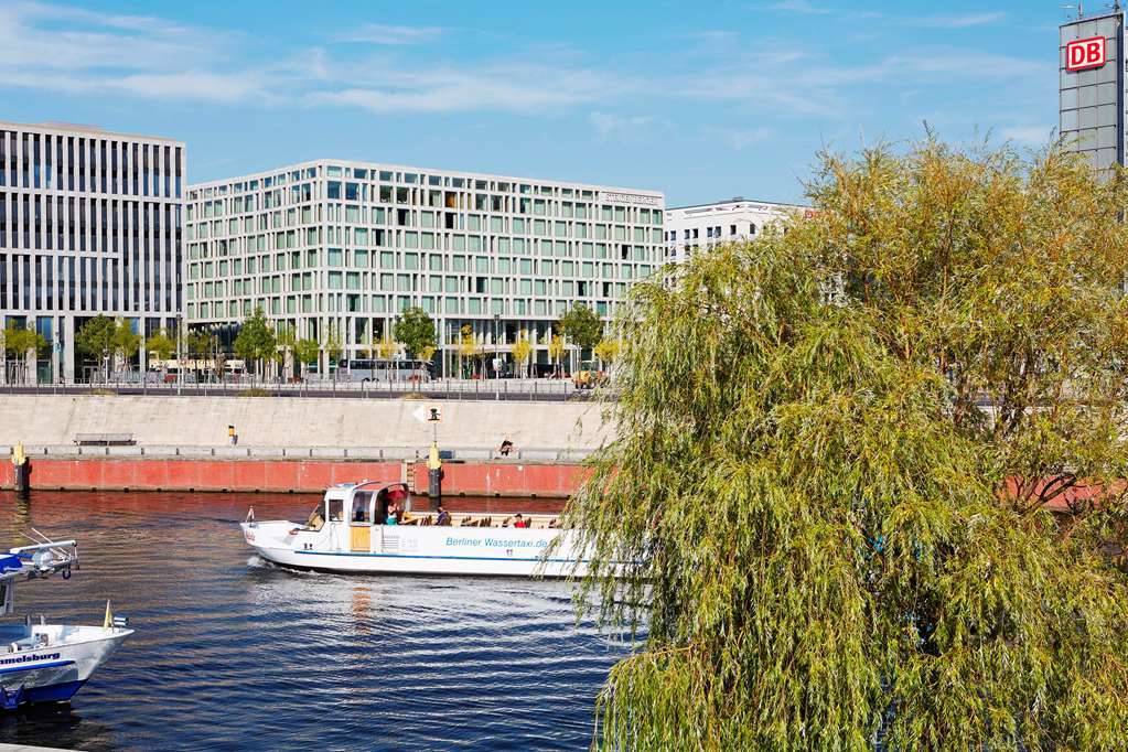 Steigenberger Hotel Am Kanzleramt Βερολίνο Εξωτερικό φωτογραφία The photo shows a scenic view of a river with a boat traveling along it. On the left side, there are smaller boats, and on the right, a large white boat is visible, likely for tourism or leisure. In the background, there are modern buildings with a s