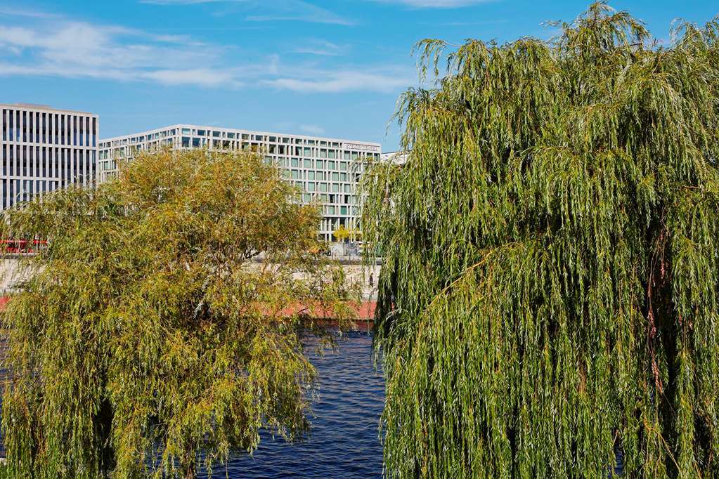 Steigenberger Hotel Am Kanzleramt Βερολίνο Εξωτερικό φωτογραφία The photo displays a scenic view featuring two willow trees with long, hanging branches. In the background, there is a body of water that reflects the sky. Behind the trees, modern buildings are visible, characterized by large glass windows. The sky 