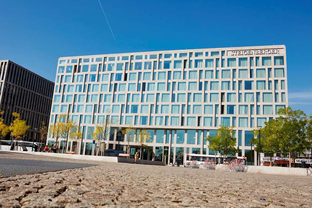 Steigenberger Hotel Am Kanzleramt Βερολίνο Εξωτερικό φωτογραφία The photo depicts a modern building with a sleek glass facade, characterized by a grid-like pattern of windows. The structure appears to be several stories tall and is set against a clear blue sky. In the foreground, there is a paved area, possibly a