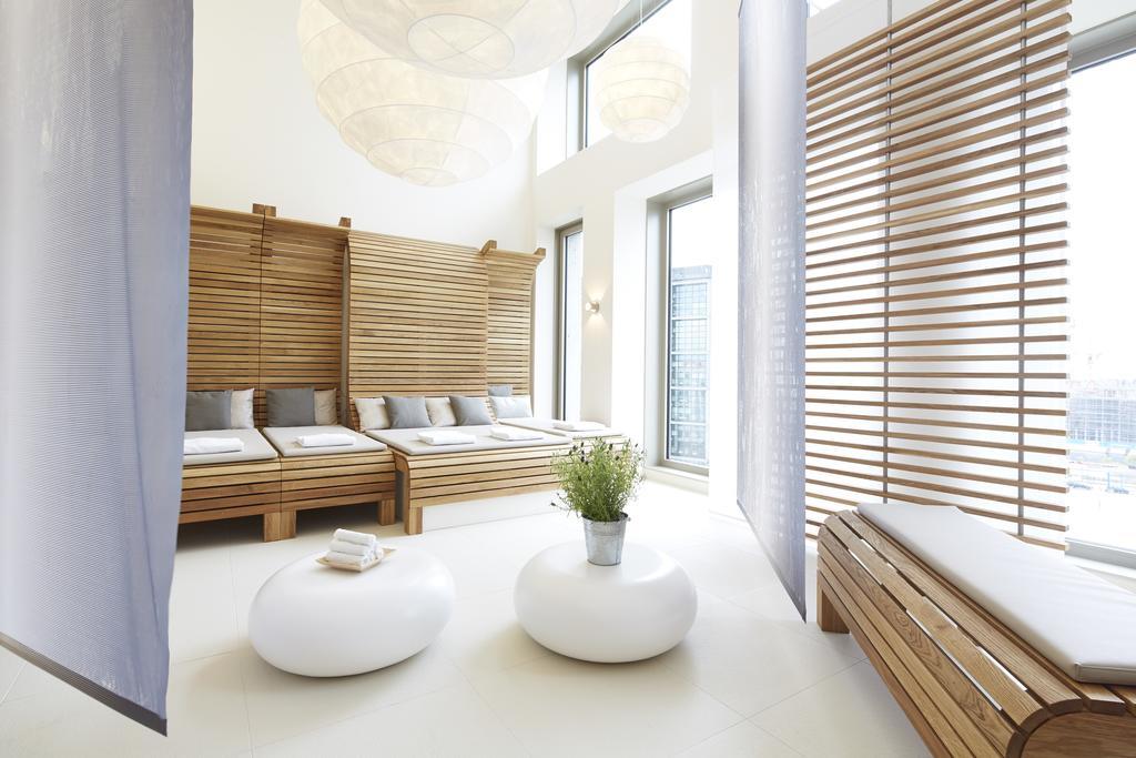 Steigenberger Hotel Am Kanzleramt Βερολίνο Εξωτερικό φωτογραφία The photo depicts a serene and modern interior space, likely a relaxation area or spa. It features wooden elements, including a slatted partition and lounge beds with soft cushions. There are round, white tables in the center, topped with a small pla