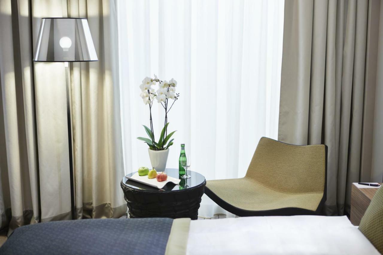 Steigenberger Hotel Am Kanzleramt Βερολίνο Εξωτερικό φωτογραφία The photo shows a cozy hotel room setting. There's a large window with sheer curtains allowing natural light to filter in. In the foreground, a small round table is accompanied by a stylish chair. On the table, there is a bottle of sparkling water an