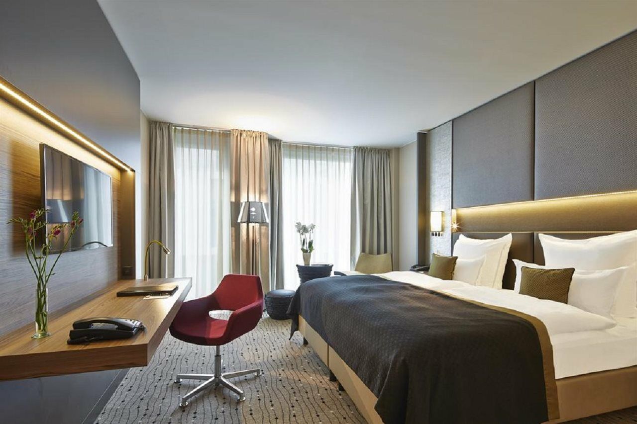 Steigenberger Hotel Am Kanzleramt Βερολίνο Εξωτερικό φωτογραφία The photo shows a modern hotel room with a clean and stylish design. It features a large bed with plush pillows and a dark bedspread. There's a wooden desk with a chair, a lamp, and a decorative vase with flowers. Natural light filters in through lar